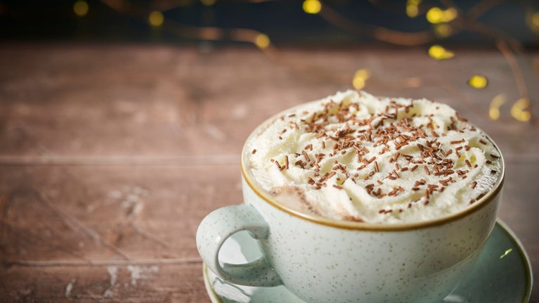 A cup of festive hot chocolate sits on a wooden table with twinkle lights